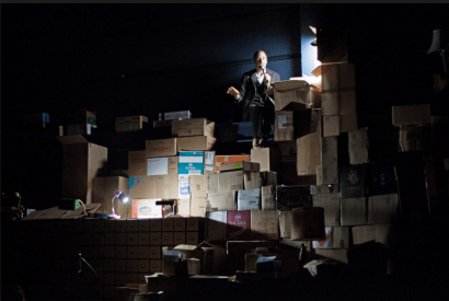 Geoff Sobelle with astonishing intimacy from the top of a mountain of boxes in 